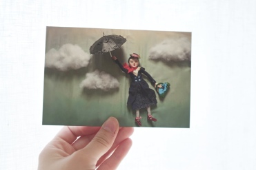 a hand holding up a postcard with an image of Mary Poppins flying through the sky with her umbrella